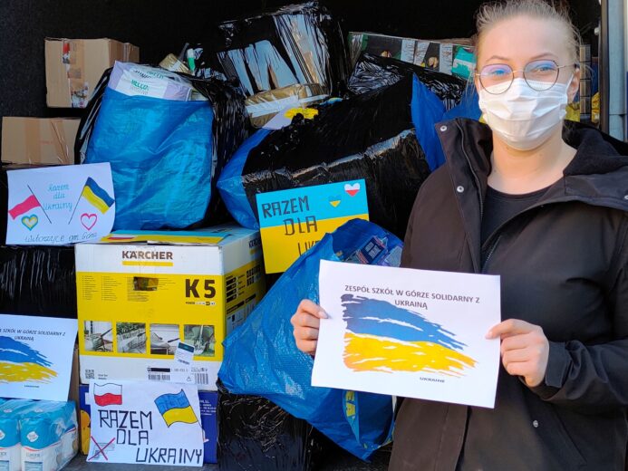 Uczennica z grafką solidarności z Ukrainą na tle załadowanego busa z darami.