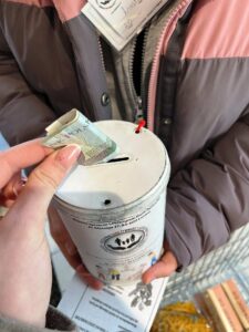 Na zdjęciu widać wolontariuszkę trzymającą puszkę akcji "Wielkanocna Zbiórka Żywności" oraz rękę osoby wpłacającej do puszki 10zł.