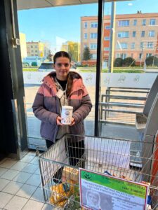 Wolontariuszka biorąca udział w akcji "Wielkanocna Zbiórka Żywności" zbiera żywność w sklepie Biedronka w Górze. Na zdjęciu widać koszyk z zebraną żywnością oraz wolontariuszkę z puszką.