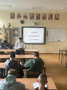 Uczeń czyta na głos książkę dla całej klasy na języku polskim. W tle na tablicy multimedialnej widnieje napis przypominający o udziale w akcji "Dzień Głośnego Czytania".