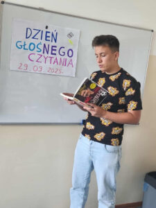 Uczeń czytający w klasie swoją ulubioną książkę na głos. W tle plakat przypominający o udziale w akcji "Dzień Głośnego Czytania" 29.09.2025 r.